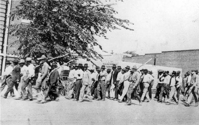 En gruppe National Guard-tropper, som bærer rifler med bajonetter festet, eskorterer ubevæpnede svarte menn til et interneringssenter etter Tulsa Race Massacre, Tulsa, Oklahoma, juni 1921.