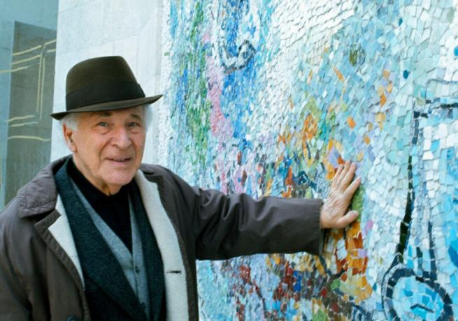 Kunstneren Marc Chagall som har hatt på seg trykker hånden mot en vegg med blå mosaikkdesign.